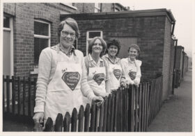 Four women dressed in aprons that each read Souper Woman looking over a fence of a back to back in the 80s.