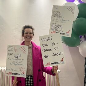 Our beaming Alice in a bright pink coat and glasses holding placards. One has "What did you think of the show?" written on in marker. The others have audience feedback like, "Fantastic performances" and "Don't underestimate the women" written on.