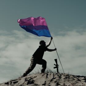 A man sticking a pink purple and blue bisexual flag in the ground on the top of a hill with a wide stance