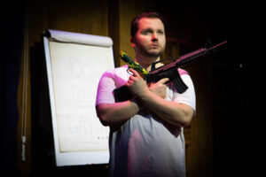 A person onstage with two toy guns across his chest with a flipchart behind.