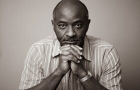 A man in a short sleeved button down shirt resting on his closed hands and starting straight down the lens.