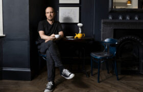 Chris Thorpe is sat crossed leg at a table, in a coffee shop, looking directly at the camera, whilst holding a white teacup. On the table is a yellow teapot, with the lid off and a white mushroom cloud escaping. Behind him are several illustrated prints on the wall.