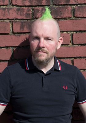 Leon with green mohican and serious expression in front of a brick wall