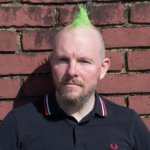 Leon with green mohican and serious expression in front of a brick wall