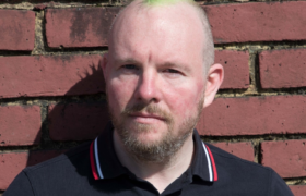 Leon with green mohican and serious expression in front of a brick wall