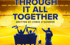 The backs of three people sat in the middle of a football stadium's pitch. Two are waving scarves and one pointing directions.