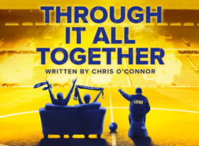 The backs of three people sat in the middle of a football stadium's pitch. Two are waving scarves and one pointing directions.