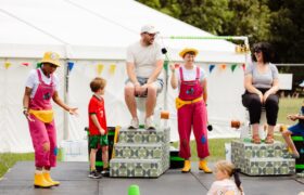 Two grownups sat on toilets on a stage with the presenters and children watching