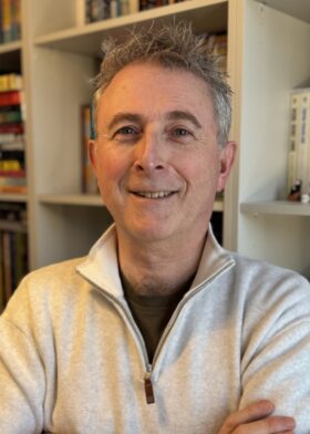 A man with grey hair stood in front of bookshelves, smiling with his arms folded.