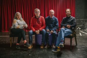 Four people sat on the edge of a stage in front of a velvet red curtain, 