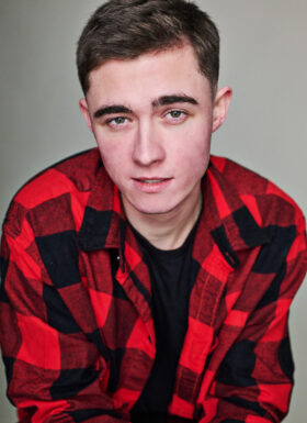 A young man in a red and black plaid shirt looking to camera