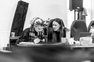 Lillie and Eija sat behind a desk in a rehearsal room in conversation as Lillie writes