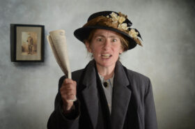 A woman in suffragette's clothing holds up a newspaper